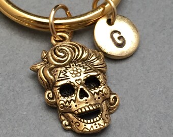 May include: Gold-toned keychain featuring a detailed sugar skull charm with a black and white design and a small round charm with the letter "G" on it.