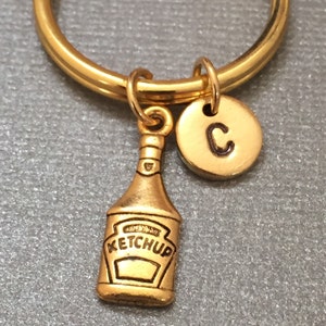 以下が含まれることがあります： 小さな金のケチャップボトルのチャームと金のチャームにCの文字が刻まれた、金のキーホルダーです。