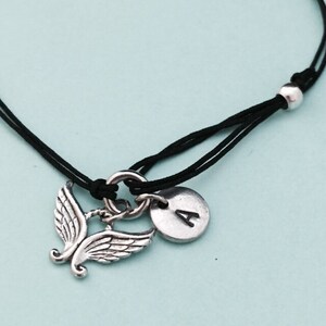 May include: A black cord bracelet with a silver angel wing charm and a silver charm with the letter A.