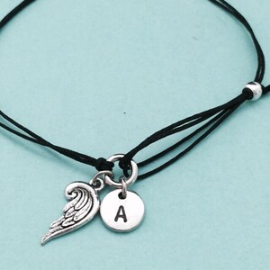 May include: A black string bracelet with a silver charm featuring an angel wing and a letter A.