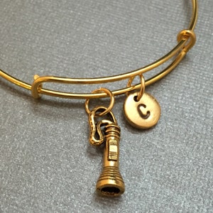 May include: A gold-toned expandable bangle bracelet with a charm of a vintage-style flashlight and a gold-toned charm with the letter 'C' on it.