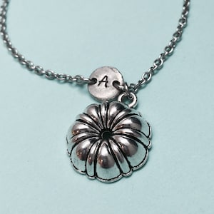 May include: A silver necklace with a small silver charm in the shape of a bundt cake pan. The charm has a small silver disc attached to it with the letter "A" engraved on it.