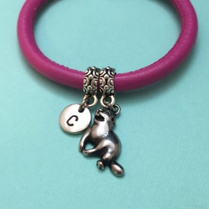 May include: A pink leather bracelet with two silver charms. One charm is a round disc with the letter "C" engraved on it. The other charm is a silver animal figurine.