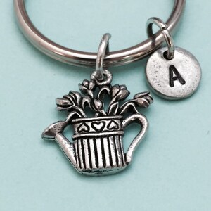 May include: Silver keychain with a watering can charm and a small silver charm with the letter A. The watering can charm has a bouquet of flowers inside.
