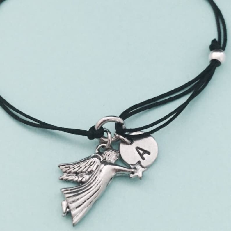 May include: A black string bracelet with a silver angel charm and a silver charm with the letter "A" on it.