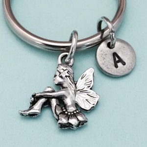 May include: Silver charm keychain featuring a sitting fairy with wings and a flower in her hair. The keychain also has a silver charm with the letter A.