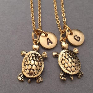 Könnte beinhalten: Zwei goldene Halsketten mit Schildkrötenanhängern und Initial-Scheibenanhängern. Die Initial-Scheiben sind mit den Buchstaben 'A' und 'G' graviert.