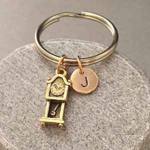 Può includere: Portachiavi dorato con un ciondolo a forma di orologio e un ciondolo con la lettera 'J'. Il portachiavi è attaccato a un anello portachiavi argentato.