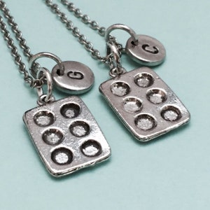 May include: Two silver necklaces with a charm of a mini muffin tin and a silver initial charm. The charms are attached to silver chains.