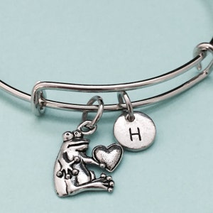 Könnte beinhalten: Silbernes Armband mit einem Frosch-Anhänger, der ein Herz-Anhänger hält, und einem silbernen Anhänger mit dem Buchstaben H.