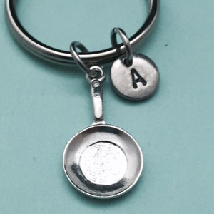 Puede incluir: Llavero de plata con un pequeño encanto de sartén y un encanto redondo con la letra A.