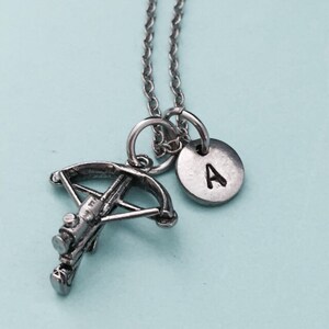 May include: Silver necklace with a crossbow charm and a small silver disc charm with the letter 'A' engraved on it.
