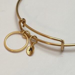 Könnte beinhalten: Ein goldenes Armreif-Armband mit einem kleinen goldenen Kreis-Anhänger und einem Blatt-Anhänger, auf dem der Buchstabe "A" eingraviert ist.