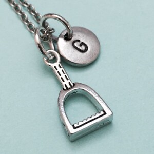 Pode incluir: Colar de prata com um pingente de estribo e um pingente de disco inicial personalizado. O pingente de disco inicial tem a letra "G" gravada.