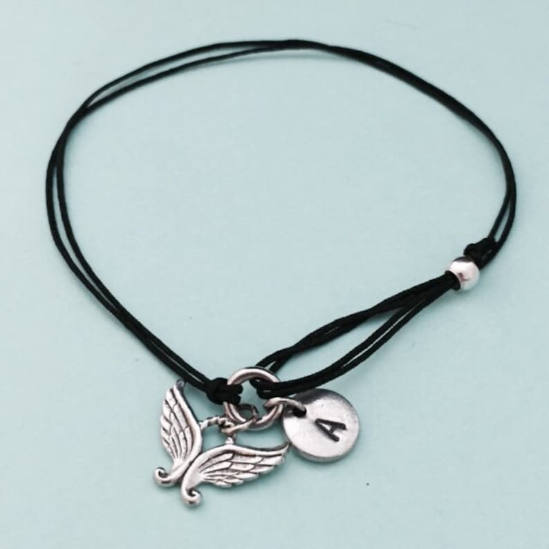 May include: A black string bracelet with a silver angel wing charm and a silver disc charm with the letter "A" on it.