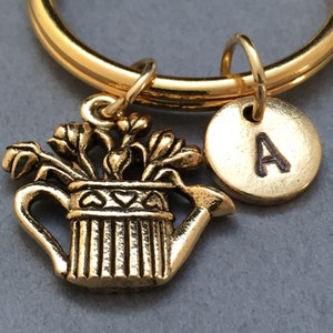 May include: Gold-toned keychain with a watering can charm and a round charm with the letter A. The watering can charm has a detailed design with flowers growing out of it.