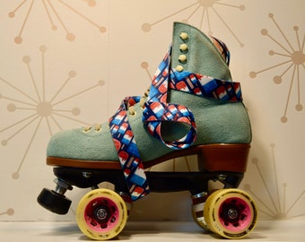 Puede incluir: Un par de patines de color azul verdoso con ruedas marrones y una correa con estampado colorido. Los patines tienen un fondo blanco con un estampado rojo, blanco y azul.