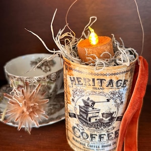 May include: A vintage-style coffee can with the text "HERITAGE BRAND COFFEE" and a graphic of a coffee grinder. The can holds a small, orange candle with a flickering LED flame, surrounded by dried twigs and moss. A decorative teacup is in the background.