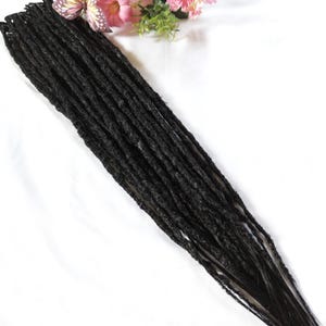May include: A bundle of dark brown dreadlocks, arranged diagonally against a white background. The dreadlocks are thick and textured, with a natural appearance. Pink flowers and a butterfly are visible in the upper right corner.