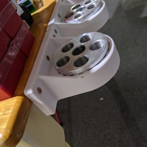 May include: A white 3D-printed wall-mounted bracket featuring a circular silver metal plate with five threaded holes. The bracket is secured with two visible screws and is designed to hold cylindrical items in a vertical or angled position.