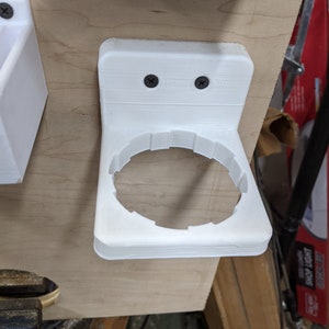 Puede incluir: Un soporte circular blanco impreso en 3D con una placa de montaje plana que cuenta con dos tornillos negros. El soporte tiene un borde interior con muescas diseñado para sostener un objeto cilíndrico, fijado a una superficie de madera clara.