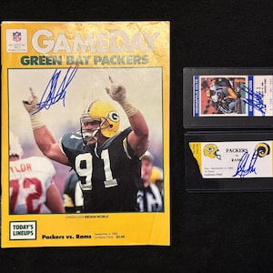 May include: A vintage Green Bay Packers Gameday magazine cover featuring a player in a green and gold uniform, with the text "Packers vs. Rams." Also includes a signed ticket and a trading card, both featuring Packers imagery.