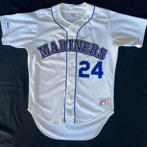 Op de afbeelding: Wit baseballshirt met blauwe en gouden bies. Het shirt heeft het woord "Mariners" in blauwe en gouden letters en het nummer "24" in blauwe letters. Het shirt is gesigneerd door de speler.
