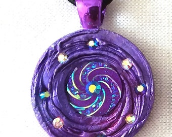 May include: A purple pendant necklace with a swirling design and iridescent center. The round pendant is adorned with small, sparkling crystals and hangs from a black cord. The pendant measures approximately 1.5 inches in diameter.