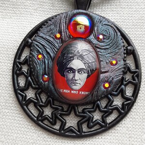 May include: Black pendant necklace with a round, black metal frame featuring a star design. The center of the pendant has a red, round, glass cabochon with a black and white image of a man in a turban with the text "The Man Who Knows".