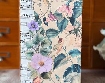 May include: A rectangular journal with a cover featuring a collage of vintage sheet music on the left side and a floral pattern with pink flowers, green leaves, and a small winged figure on the right. A pink fabric ribbon extends from the top.