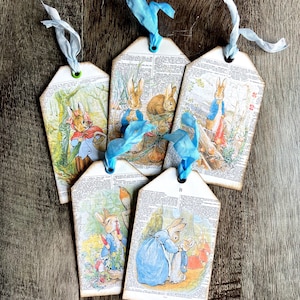 May include: Set of five vintage-style gift tags featuring illustrations of rabbits in various scenes. Each tag is made from book pages, with a blue ribbon attached. The tags have a rustic, aged appearance.