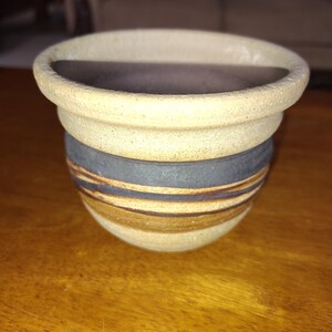 May include: A handmade ceramic bowl with a layered design. The top section is a light beige color, followed by a dark gray band, and a lower section with brown and tan stripes. The bowl has a rounded shape and a wide opening.