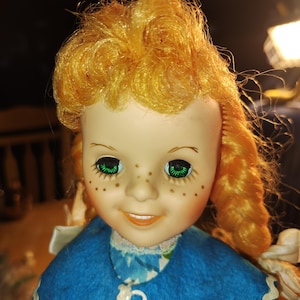 May include: A vintage doll with bright green eyes and freckles. The doll has orange hair styled in curls and braids. She wears a blue dress with a white lace collar and a decorative white trim. The doll's expression is a slight smile.