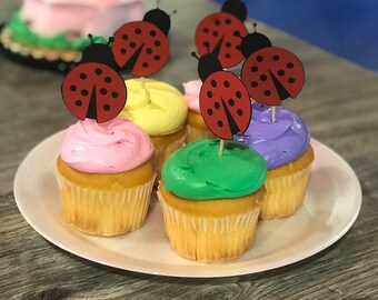 May include: Four cupcakes decorated with colorful frosting and ladybug toppers. The cupcakes are on a white plate.
