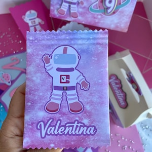 May include: A party favor bag with a purple and pink galaxy design, featuring an astronaut illustration and the name "Valentina" in cursive. Other boxes with astronaut and planet graphics are visible.