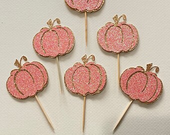 May include: Six pink glitter pumpkin cupcake toppers with gold glitter accents. The toppers are attached to wooden skewers.