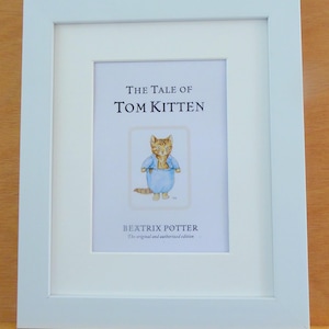 May include: A white rectangular picture frame containing a print of The Tale of Tom Kitten by Beatrix Potter. The print features a central illustration of a ginger kitten wearing blue clothing, with the title and author name printed in black text.
