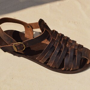 May include: A single brown leather sandal with a woven strap design. The sandal features a buckle closure and a strap around the ankle. The sandal is displayed on a sandy surface.