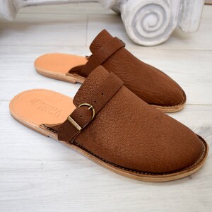 May include: A pair of brown leather slip-on shoes with a buckle strap. The shoes have a light brown sole and are made of a textured leather.