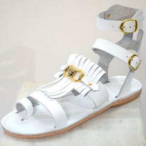 May include: White leather sandal with a toe loop and ankle strap. The sandal features gold-colored buckles and decorative elements, including a fringed accent. The sole is a light brown color.