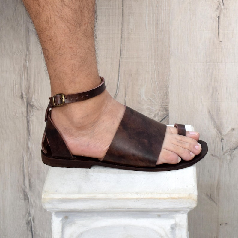 May include: Brown leather sandal with a toe loop and ankle strap, featuring a buckle closure. The sandal is worn on a foot, showcasing the design and craftsmanship. The footwear is designed for comfort and style.