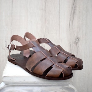 May include: A pair of brown leather sandals with a cage-like design. The sandals have a flat sole and a strap that buckles around the ankle. The leather is a medium brown color, and the sandals appear to be new.