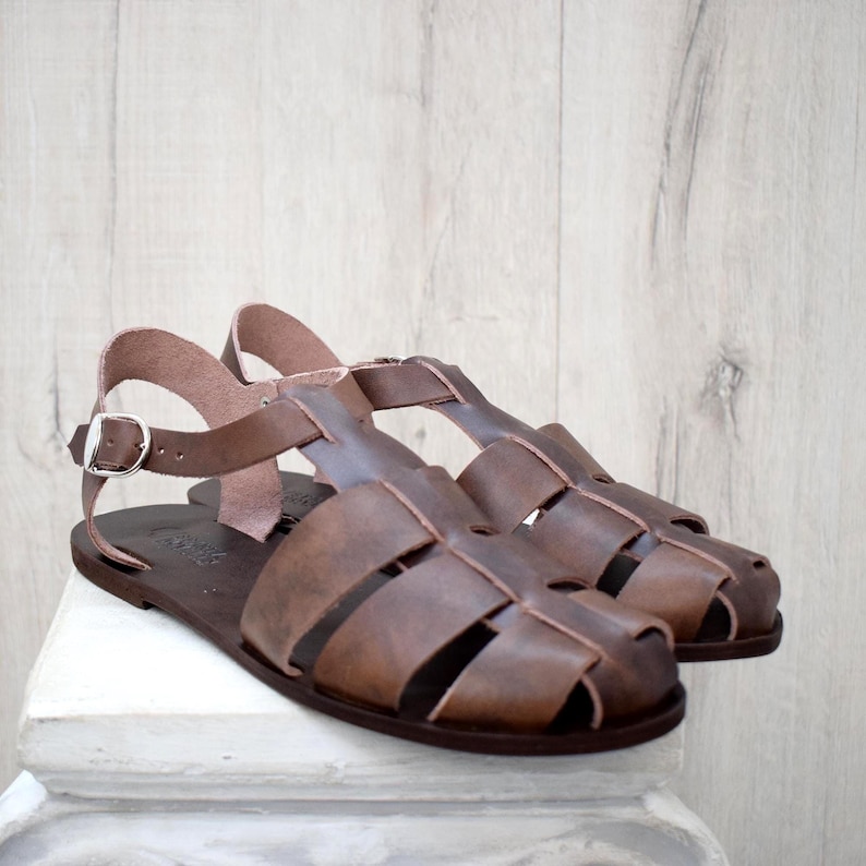 May include: A pair of brown leather sandals with a cage-like design. The sandals have a flat sole and a strap that buckles around the ankle. The leather is a medium brown color, and the sandals appear to be new.