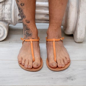 May include: Orange leather sandals with a T-strap design. The sandals have a simple, minimalist style with a buckle closure around the ankle. The soles are a light brown color.