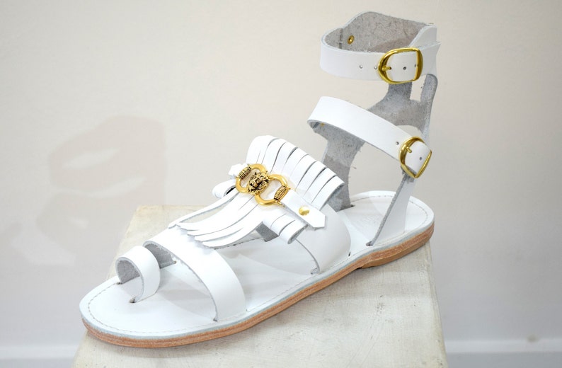 May include: White leather sandals with a toe loop and ankle strap. The sandals feature gold-colored buckles and a decorative element with fringe and a gold-colored emblem. The sole is a light brown color.