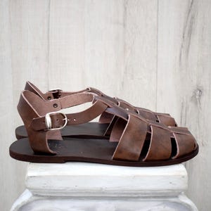 May include: Brown leather sandals with a cage-like design. The sandals have a buckle closure and a flat sole. The leather appears to be distressed, giving them a vintage look. The sandals are displayed on a white surface against a light wood background.