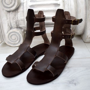 May include: A pair of brown leather gladiator sandals. The sandals have multiple straps and buckles, with a high ankle design. The leather appears to be a rich, dark brown color. The sandals are displayed on a light-colored surface.