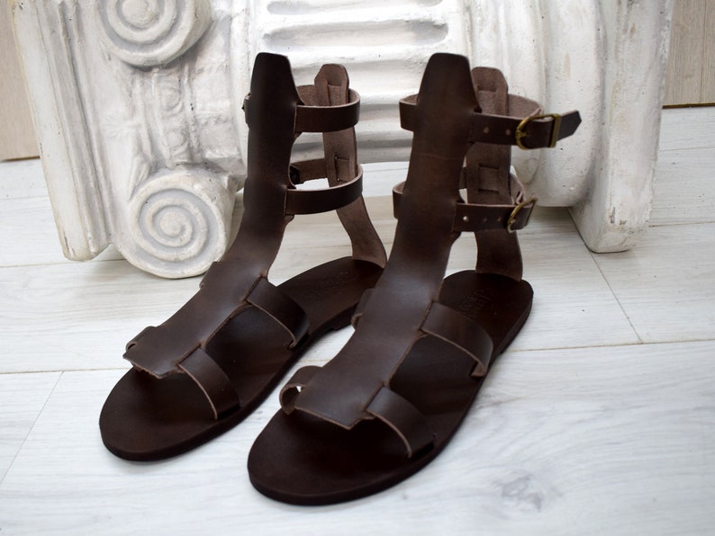 May include: A pair of brown leather gladiator sandals. The sandals have multiple straps and buckles, with a high ankle design. The leather appears to be a rich, dark brown color. The sandals are displayed on a light-colored surface.