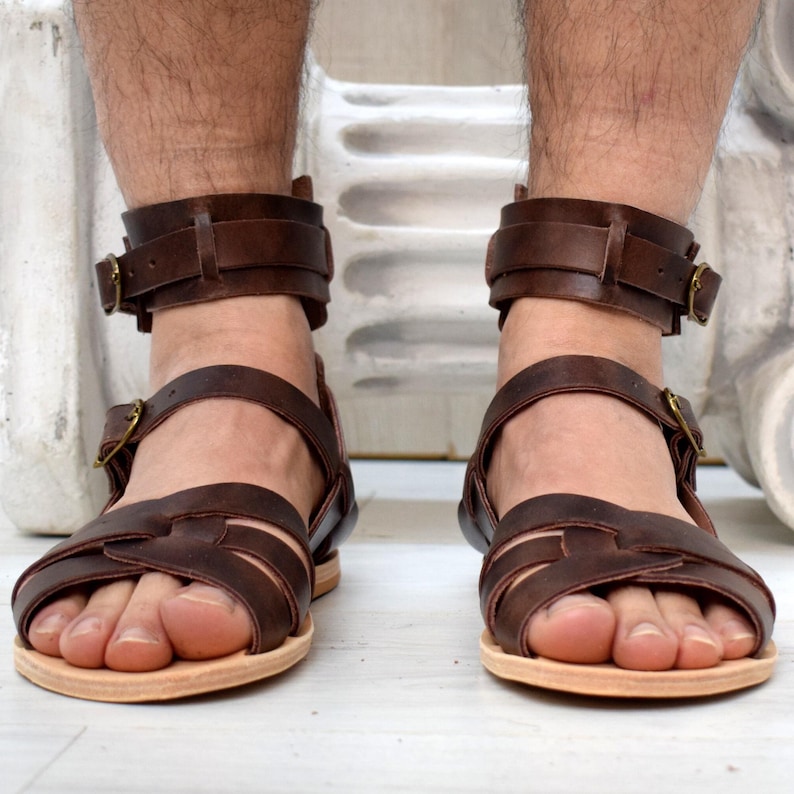 May include: Brown leather sandals with multiple straps and ankle straps secured with gold-colored buckles. The sandals have a light-colored sole. The image shows the sandals on a person's feet.