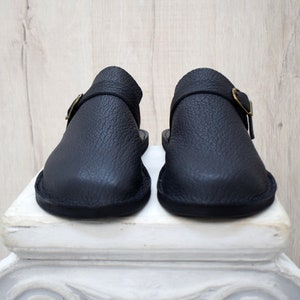 May include: A pair of black leather clogs with a textured finish. Each clog features a strap with a gold-colored buckle. The shoes are displayed on a white pedestal against a light wood background.